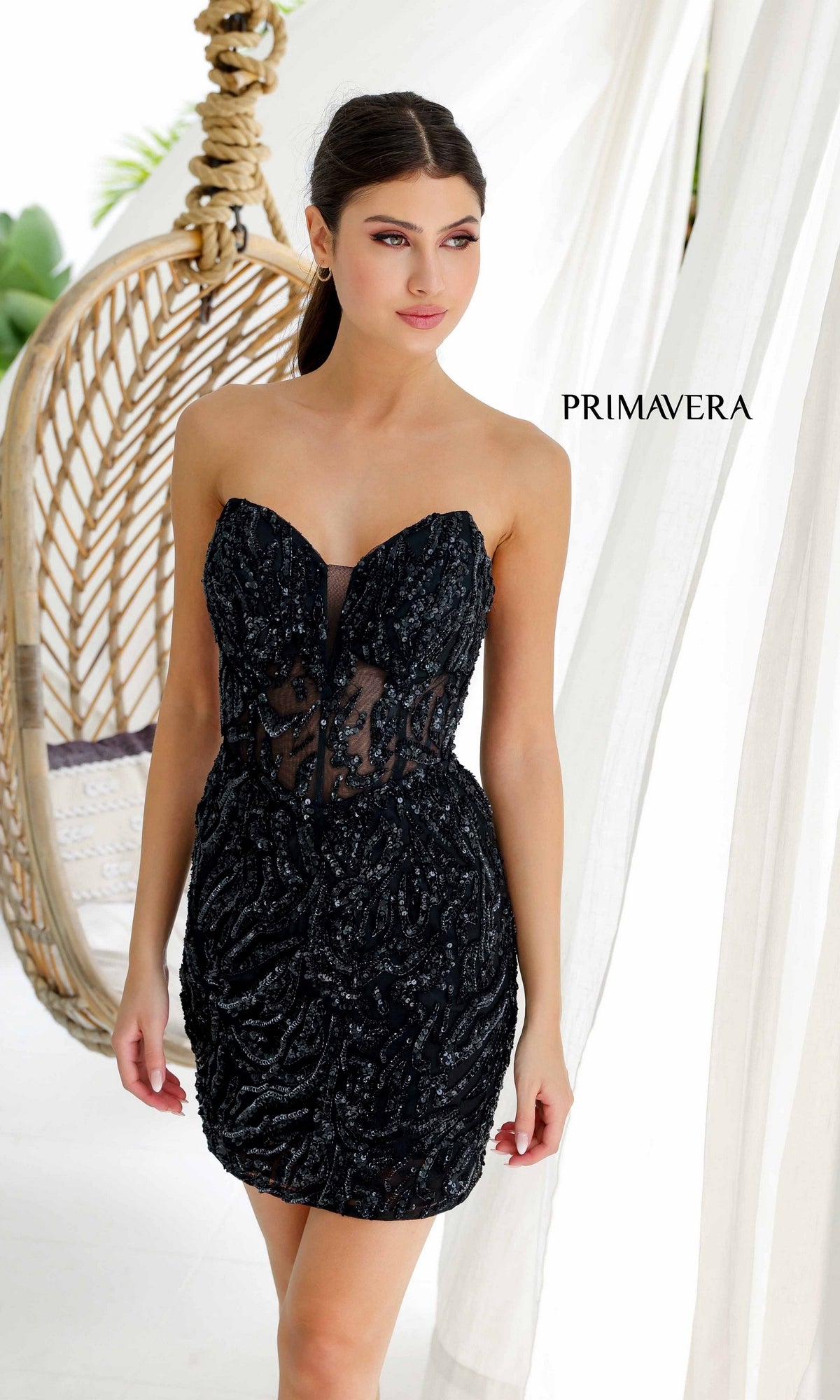 A woman stands indoors near a hanging chair and white curtains, wearing the Short Homecoming Dress 4222 by Primavera—a sleek, black sequined strapless mini dress. The word